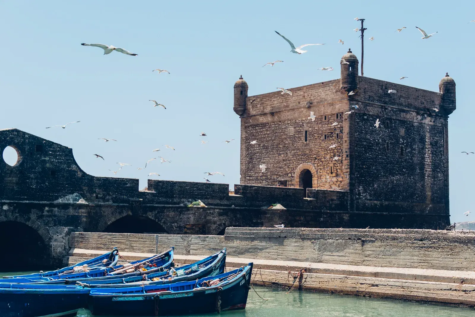 The stone citadel of Essaouira harbour with seagulls overhead and blue fishing boats below